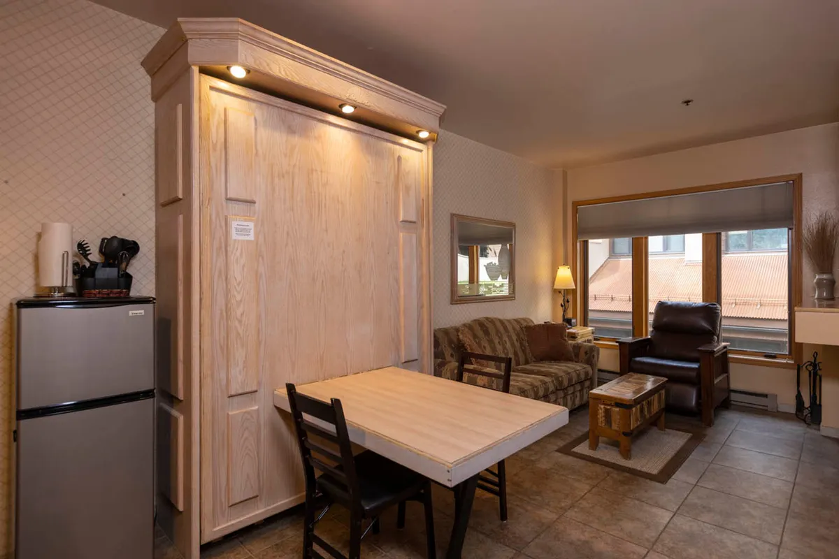 A warm, inviting condo interior featuring a neatly tucked Murphy bed, a small dining table set for two, a plush armchair, and a window that frames sunset views over the mountains. The space feels cozy and perfect for relaxing after a day in the Colorado outdoors.