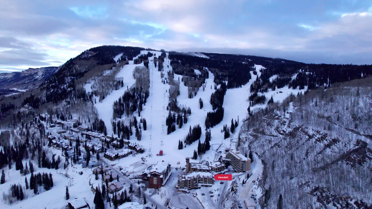 A sweeping view of the resort buildings nestled at the base of snow-covered ski slopes, surrounded by tall pine trees and set against a dramatic mountain backdrop. The scene captures both the natural beauty and lively atmosphere of the village, with guests visible enjoying winter activities and relaxing by the lodges.