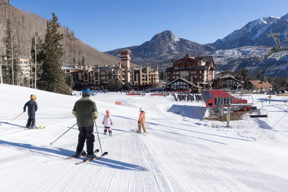 A lively scene featuring families and skiers on the slopes with resort buildings nestled at the base of a majestic mountain backdrop. The ski lift and mingling groups evoke a sense of community and fun, while the cheerful atmosphere captures the excitement of a family-friendly resort environment.