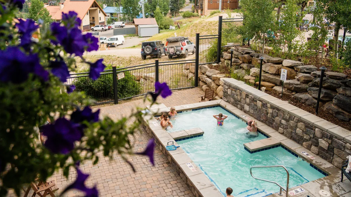 A serene resort amenities area with a bubbling hot tub and heated swimming pool, surrounded by flowering plants and set against a backdrop of mountains. The space is welcoming, with guests relaxing and socializing while enjoying sunset views.