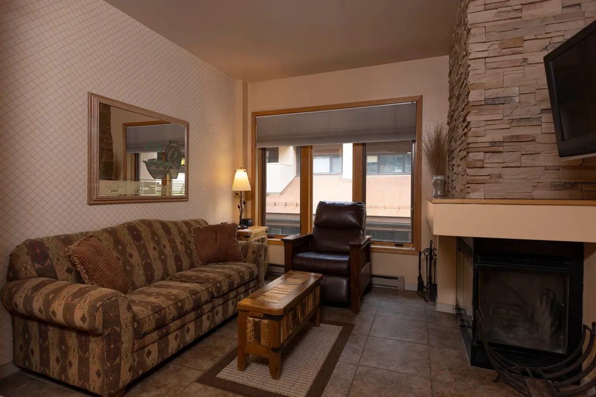 A warm, inviting living room with a plush sofa and armchair arranged around a wood-burning fireplace. Sunlight streams in through a large window, illuminating a coffee table and creating a cozy, welcoming ambiance ideal for relaxing, socializing, or unwinding after a day on the slopes.
