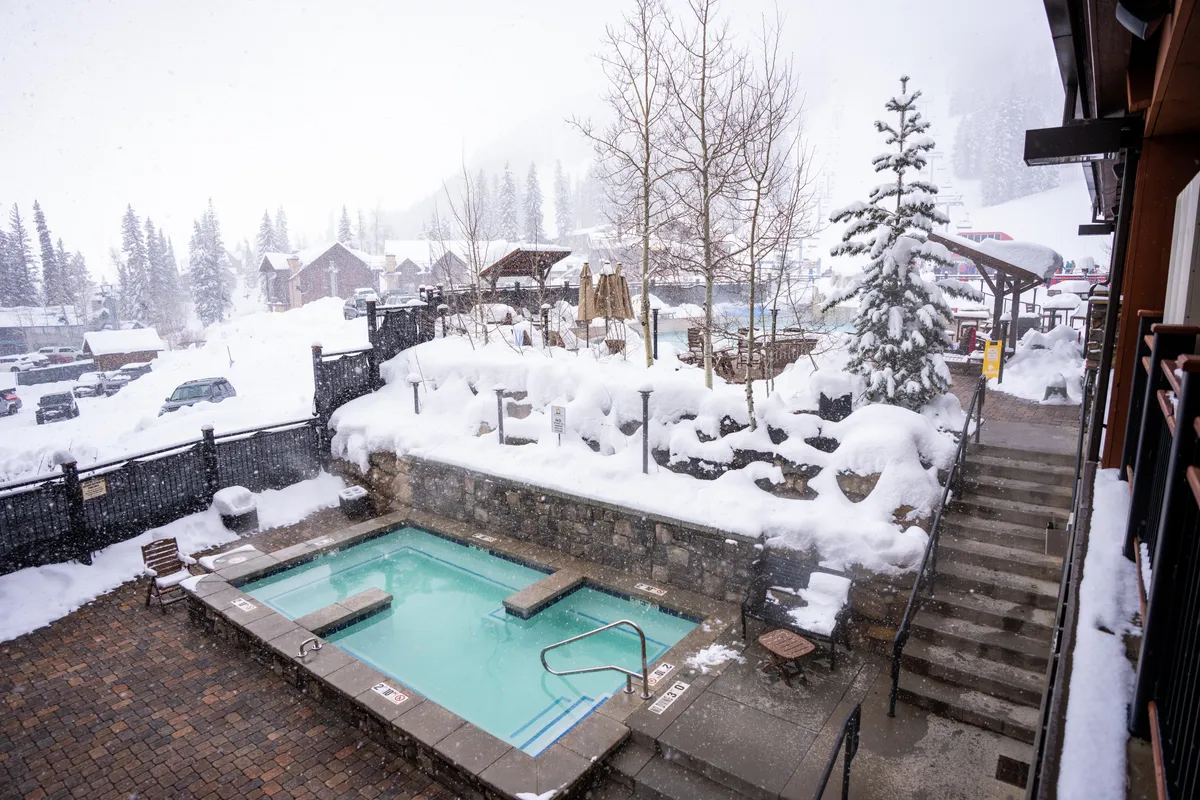 A rooftop hot tub surrounded by snow-covered landscape and trees, with a staircase leading to the amenity. The atmosphere is tranquil and inviting, with steam rising from the hot tub and hints of warmth contrasting the chilly, wintry setting.