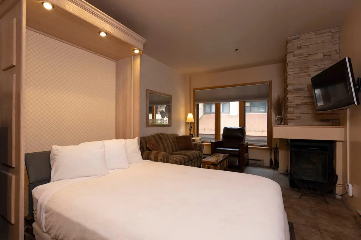 A cozy nook in the living area with a bed, a crackling fireplace, a comfortable seating area, and a window bringing in soft natural light. The space suggests a blend of relaxation possibilities—reading, watching TV, or simply enjoying the ambiance.