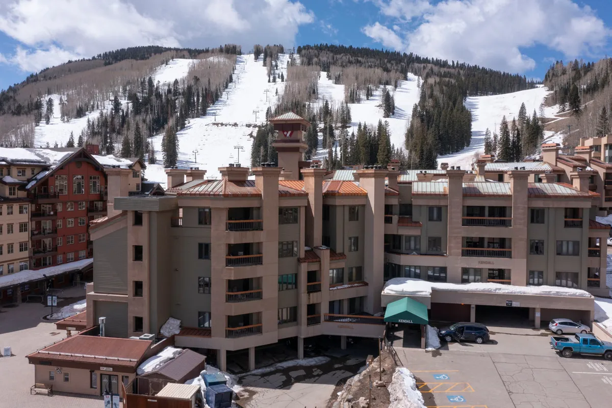 A striking exterior of the resort building framed by snow-covered mountains, with nearby ski slopes clearly visible. The scene captures the proximity to winter action, showcasing skiers and snowboarders enjoying the landscape. Architectural details of the lodge contribute to an inviting, upscale atmosphere.