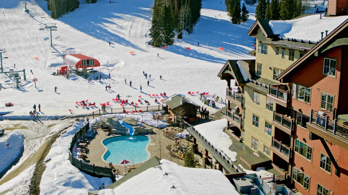 An energetic winter scene featuring an outdoor heated pool, ski slopes, a ski lift, a lodge building, and people skiing. The pool is set amidst the resort's lively environment, emphasizing both active and relaxing experiences.